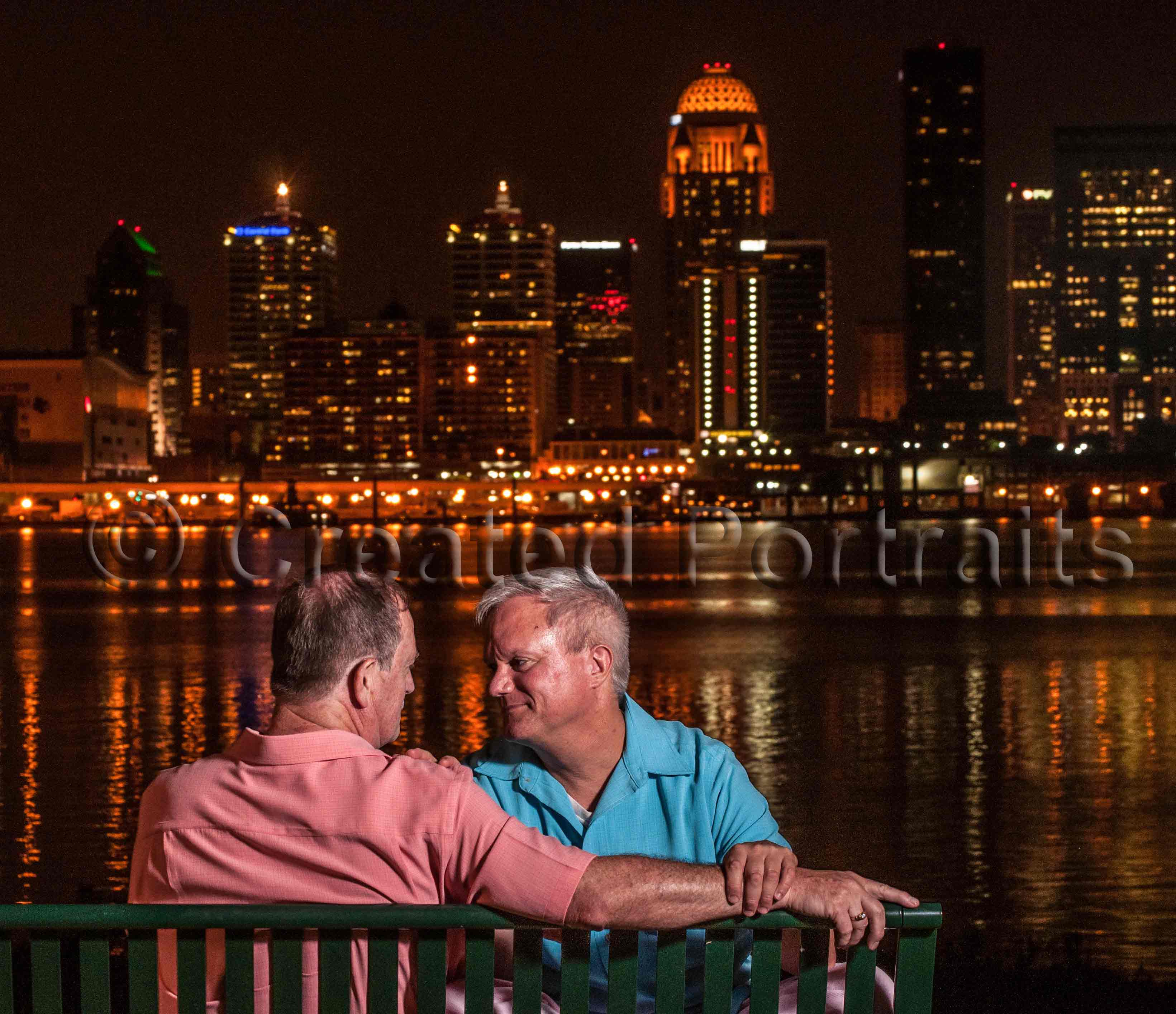 gay couple sitting on a park bench at night with Ohio river and city in the background.